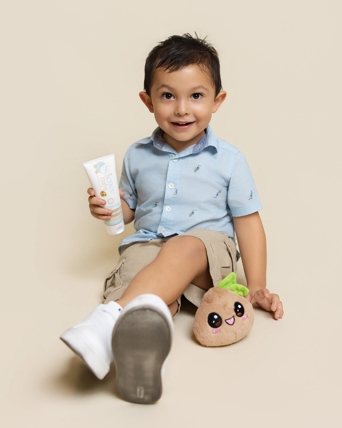A young boy sitting on a beige studio floor wearing a light blue short-sleeve button-up shirt, denim shorts, and white sneakers, holding a Science Baby Body Lotion. A small plush character toy rests beside him.
