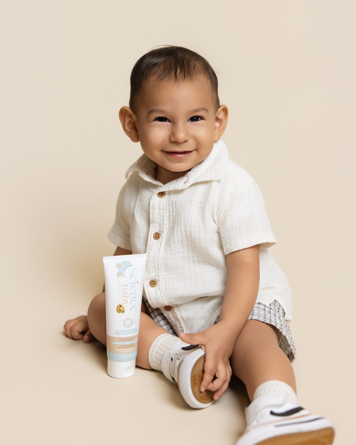 A baby sitting on a beige studio floor wearing a white short-sleeve button-up shirt and plaid shorts, holding a tube of Science baby lotion next to them.
