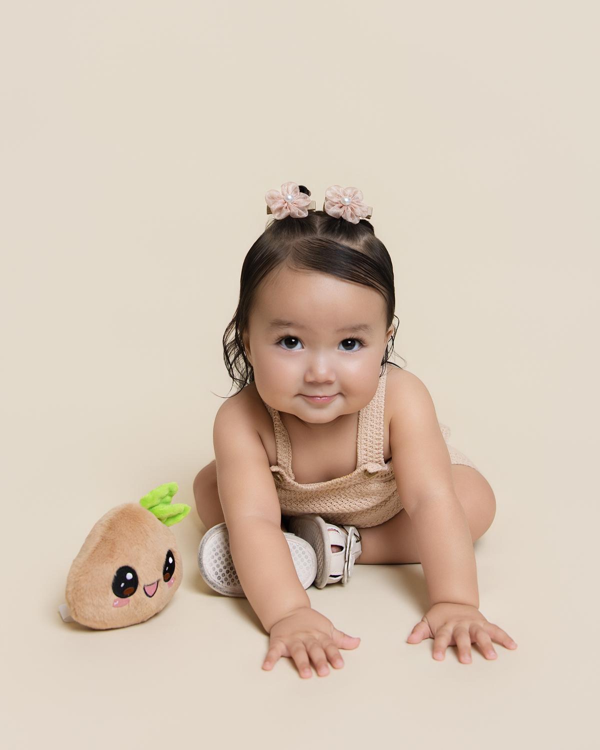 A baby girl sitting on a beige studio floor, wearing a beige knit sleeveless romper, white and gray sneakers, and a pink flower headband. A small plush toy is next to her.