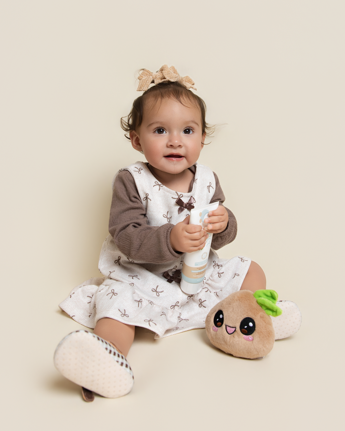 A baby girl sitting on a beige studio background, wearing a brown long-sleeve top and a white pinafore dress with brown butterfly prints, holding a Science Baby Body Lotion, with a plush toy next to her.