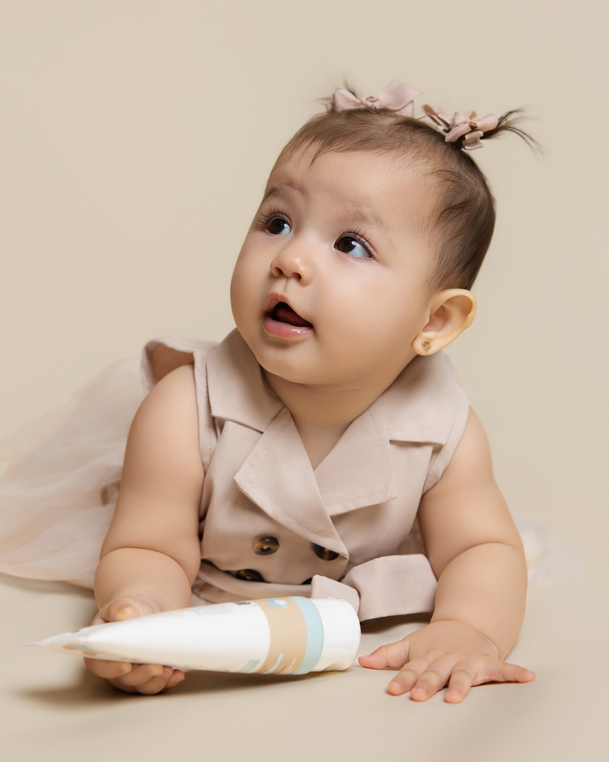 A baby girl lying on her stomach, wearing a beige sleeveless double-breasted vest and a sheer tulle skirt, holding a Science Baby Body Lotion. She has a small pink bow in her hair.