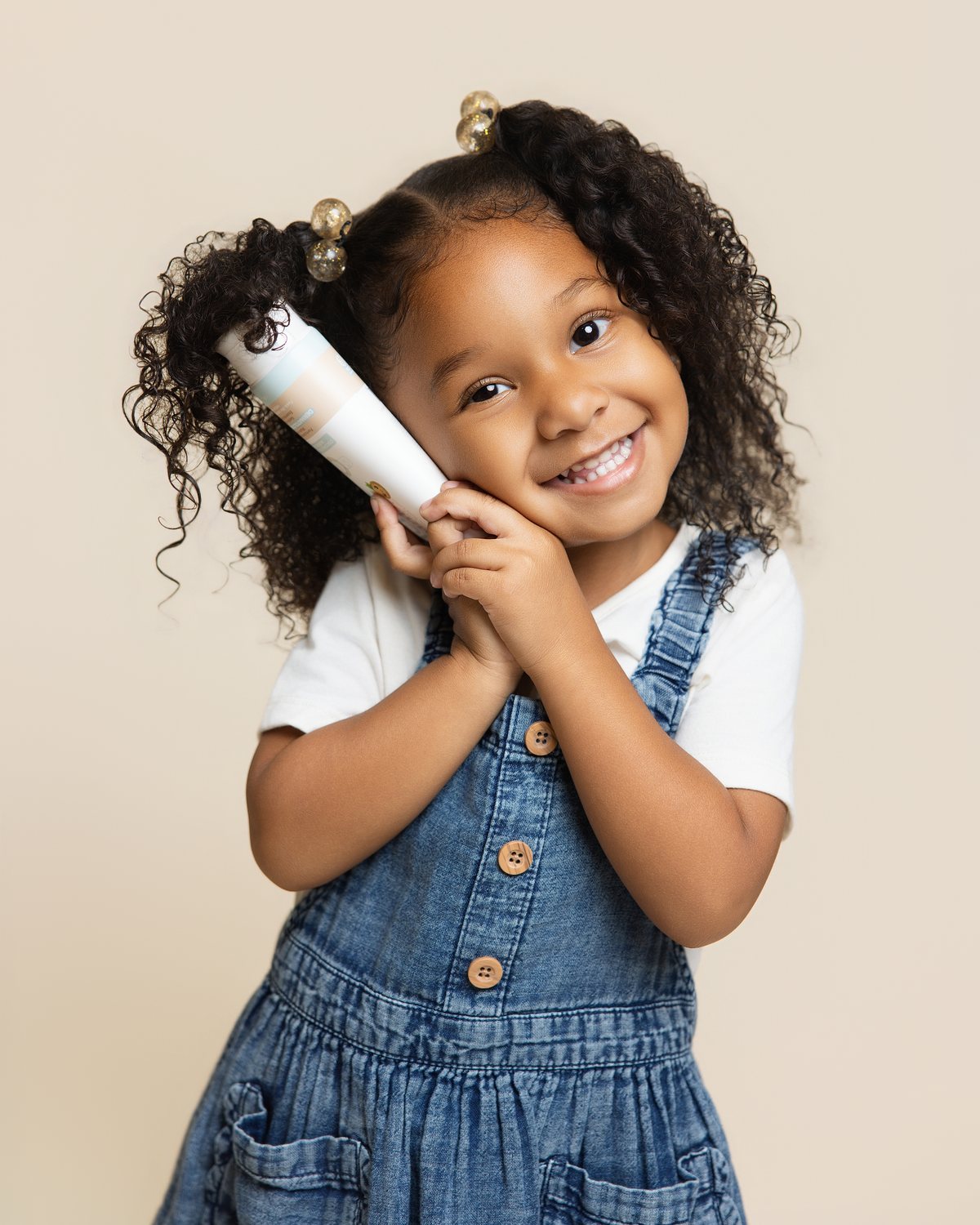 A young girl with curly hair wearing denim overalls and a white t-shirt holds a Science Baby Body Lotion near her face, posing against a solid beige background.