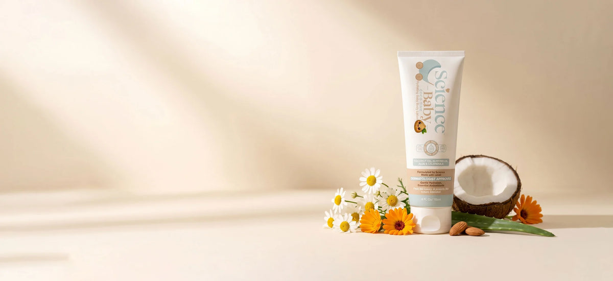 A tube of Science Baby lotion stands on a beige surface with decorative flowers, a halved coconut, and almonds. The background is a soft warm beige with light shadows.